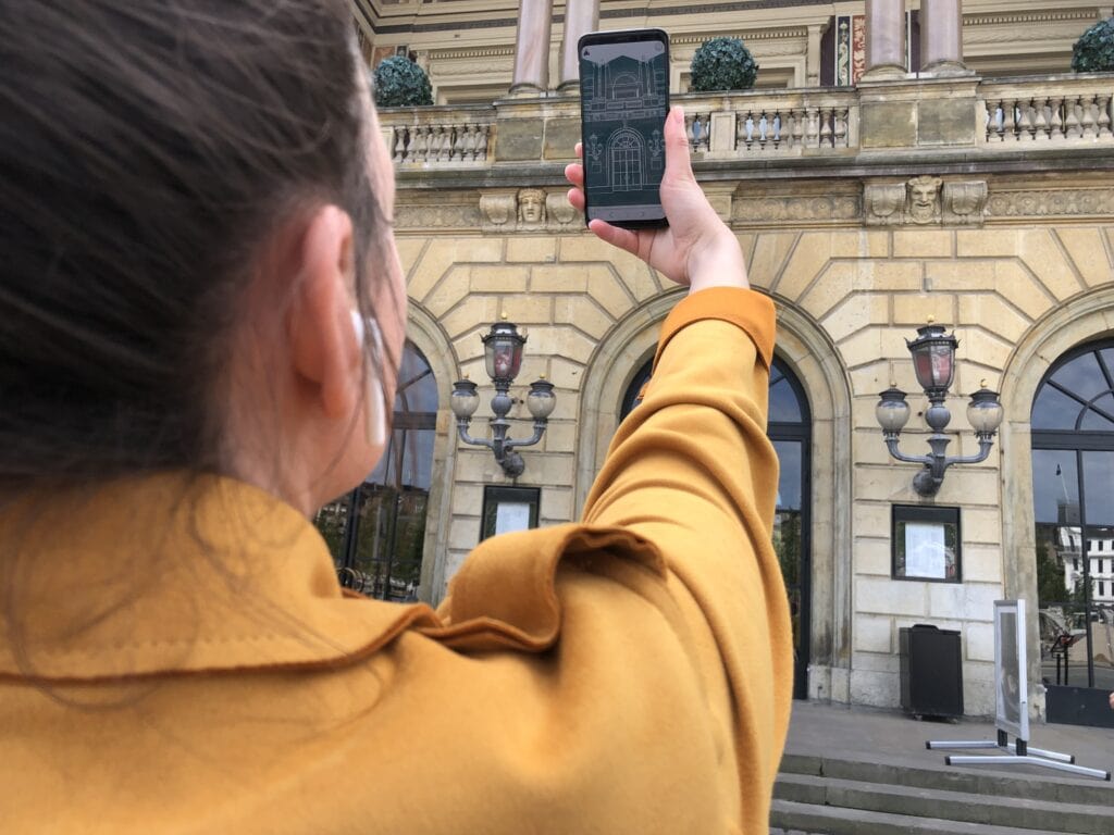A woman in a yellow coat is holding up her smartphone, on the screen you can see a sketch of the building she is standing in front of
