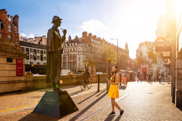A young woman in a yellow dress is standing in front of the Sherlock Holmes statue on Baker Street