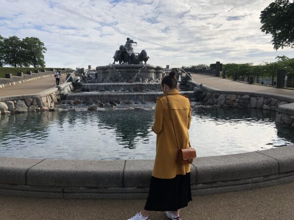 A young woman wearing a yellow coat is standing in front of the Gefion fountain in Copenhagen