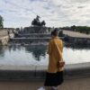 A young woman wearing a yellow coat is standing in front of the Gefion fountain in Copenhagen