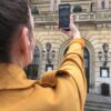 A woman in a yelow coat is atnding in front of the Danish Royal theatre. She is holding up her smartphone