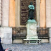 A woman in a blue coat is looking up at a bronze statue of a man